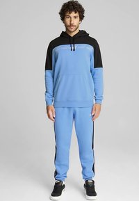 Blue hoodie with black accents; features a front pocket. Matching blue pants with side black stripes. Smooth material, athletic fit, and logo detail.