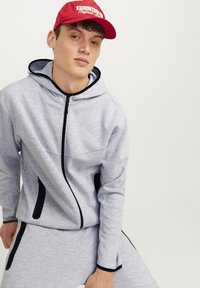 Sivi zip-up dukserica s crnim akcentima i kapuljačom sa vezicom, uparena sa odgovarajućim sivoj hlačama, nošena sa crvenom kapom sa bijelim natpisom.