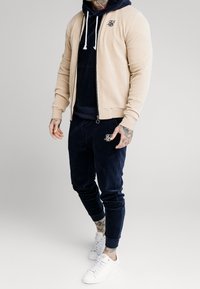 Giacca beige in corda con zip sopra un hoodie in corda blu navy e pantaloni coordinating, con logo e sneakers bianche. Texture morbida in tutto.