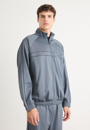 Grå zip-up treningsjakke med høy krage, elastisk midje og kontrasterende sømmer. Glatt, lett stoff som er egnet for treningstøy.