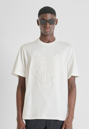 Young man wearing black sunglasses and a white t-shirt with an embossed tiger face design, standing against a plain background.