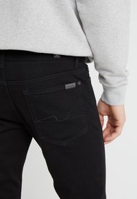Jeans de mezclilla negros con corte ajustado, con un bolsillo trasero con una etiqueta y un sutil detalle de costura. Combinados con un sudadera gris claro.