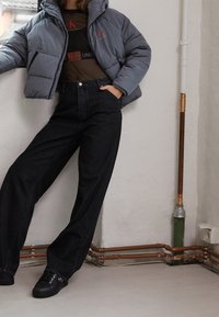 Szara kurtka puchowa z kapturem, czarny t-shirt z logo, czarne dżinsy z wysokim stanem i szerokimi nogawkami oraz czarne sneakersy w wnętrzu.