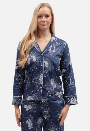 Vrouw met lang blond haar die marineblauwe pyjama met knopen draagt met witte sterrenbeelden en sterpatronen.
