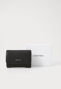 Schwarze Lederbörse mit gefaltetem Design, silbernes Calvin Klein-Logo auf der Vorderseite, begleitet von einer weißen Markenbox.