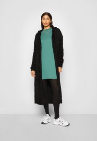 Un long cardigan noir duveteux sur une robe teal, associé à des collants noirs et des baskets blanches. Un style simple avec une coupe décontractée.