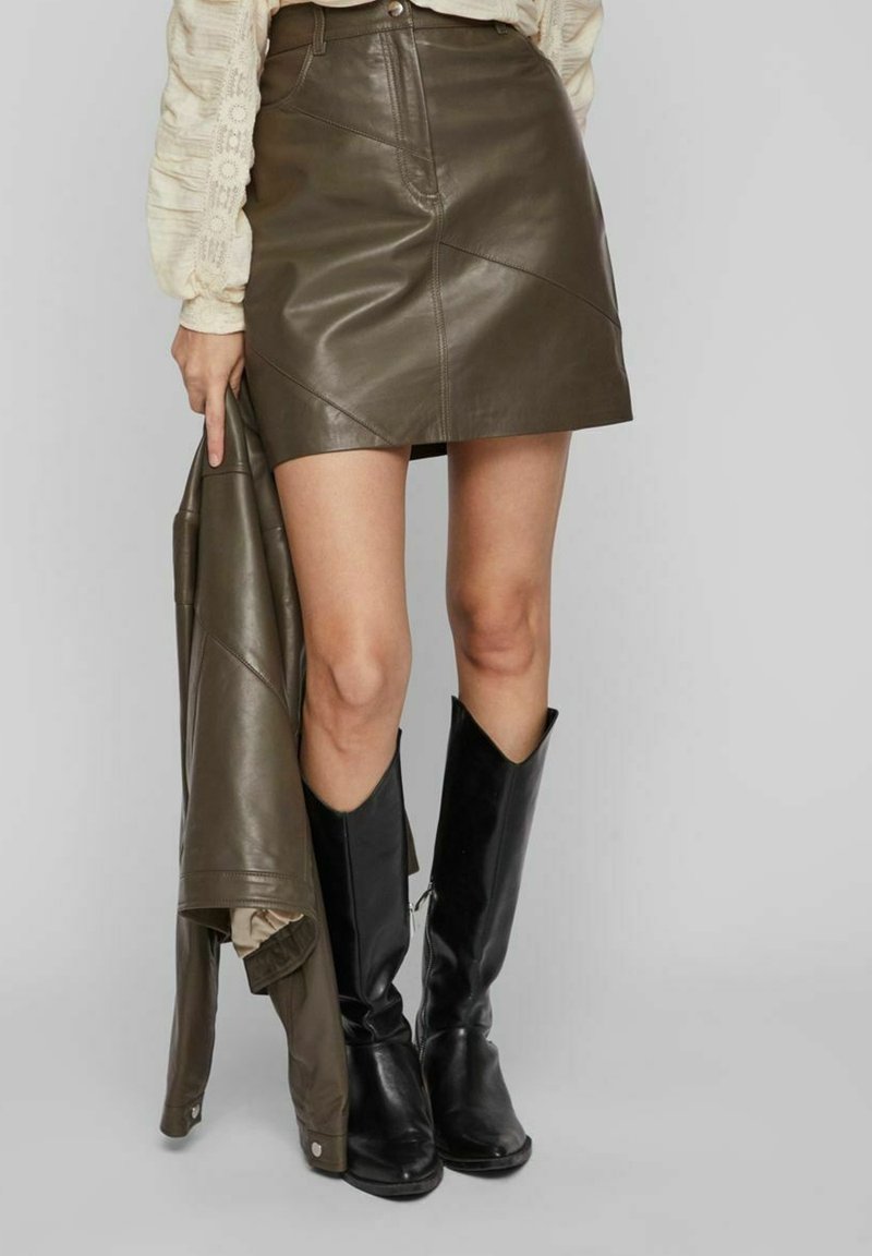Mini jupe en cuir marron avec des coutures diagonales, accompagnée d'une veste en cuir assortie portée sur un bras, et des bottes noires jusqu'aux genoux.