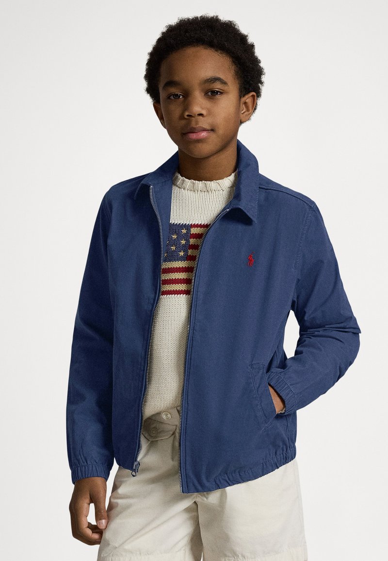 Tamsiai mėlyna lengva striukė su užtrauktuku, elastingomis rankogaliais ir šoninėmis kišenėmis. Dėvima ant kremine spalvos megztinio su raudona ir mėlyna raštu.