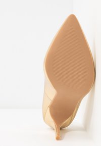 Chaussure à talon haut beige à bout pointu vue de la semelle, affichant une semelle en caoutchouc texturée et un talon fin sur fond blanc.