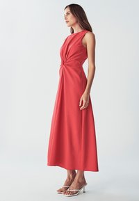 Červené midi šaty bez rukávov s prekrúteným predným detailom, vyrobené z hladkej látky. V kombinácii s bielymi strappy sandálmi na vysokom opätku.