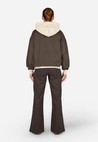 Hnedá mikina s kapucňou a krémovou podšívkou, uvoľnený strih, elastické manžety, kombinovaná s rozšírenými tmavými nohavicami. Hladká textúra a minimalistický dizajn.