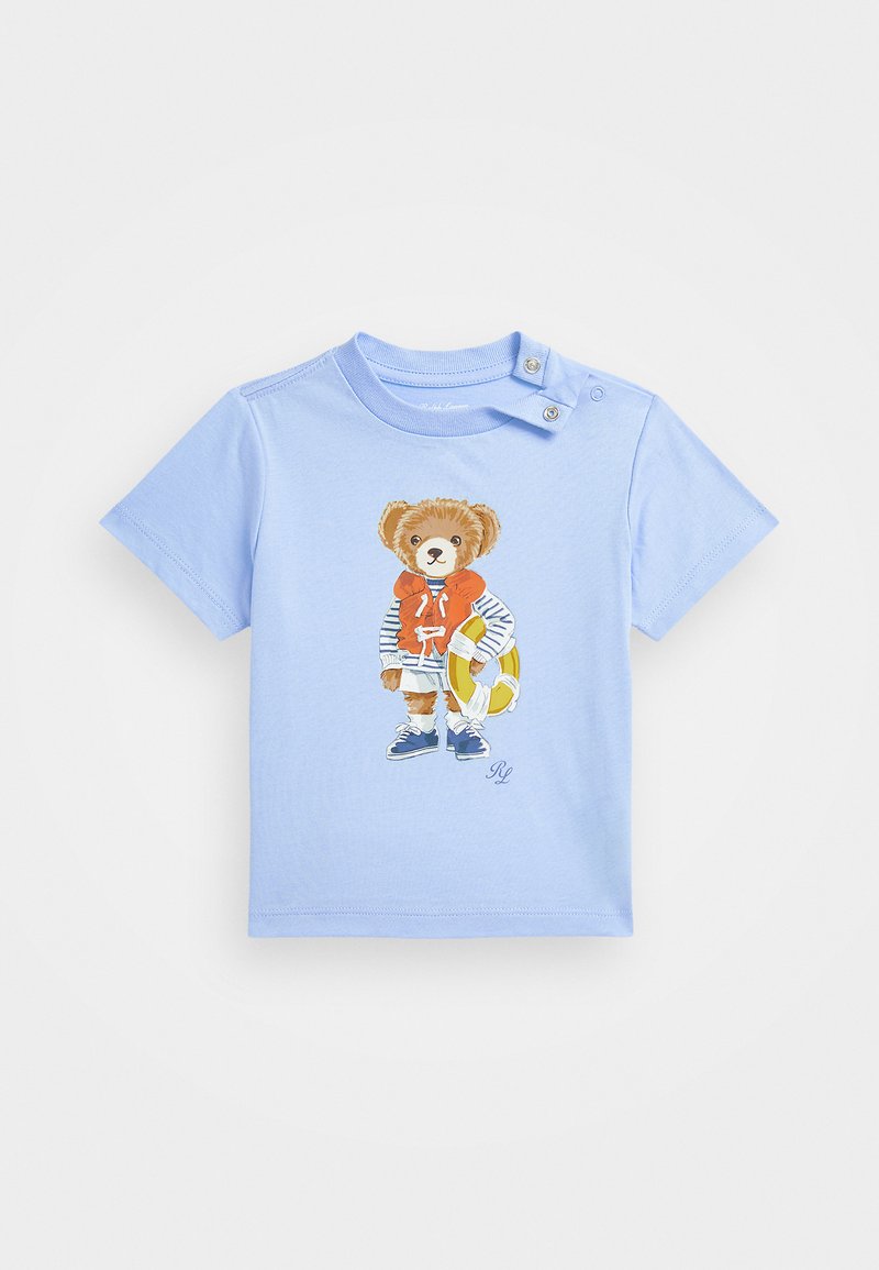 Ljusblå t-shirt med kort ärm som har en björn-grafik i en orange huvtröja och randig skjorta, som håller i en livboj. Tryckknappar på axeln.