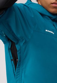 Türkisfarbene wasserdichte Jacke mit Kapuze, ausgestattet mit seitlichen Belüftungsreißverschlüssen und dem Markennamen "Mammut" in Weiß aufgedruckt. Glatte Textur.