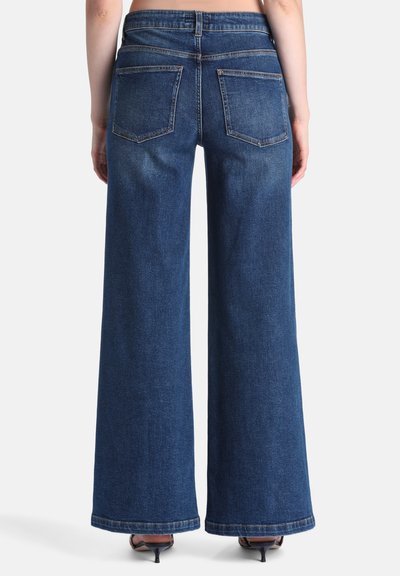 Personne portant un jean bleu foncé à jambes larges, debout de dos, avec des poches arrière visibles et des sandales à talons moyens sur un fond clair uni.