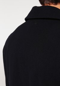 Dunkelblauer Wollmantel mit weitem Kragen und einem dezenten Logodetail auf der Rückseite. Weiche Textur, glatte Oberfläche.