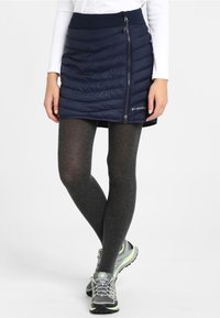 Marinblå isolerad kjol med quiltat mönster, som har en sidogylf och logotypdetalj. Matchad med mörka strumpbyxor och grå sportskor.