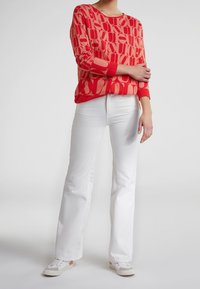 Jersey rojo con patrón, de manga larga y escote redondo, combinado con vaqueros acampanados blancos y zapatillas de color claro.