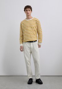 Tânăr în picioare, îmbrăcat cu o cămașă cu dungi galbene și albe, pantaloni albi cu curea neagră și pantofi negri cu șiret.