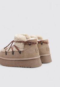 Bottines en daim beige avec une doublure beige moelleuse, une semelle en caoutchouc texturée et des lacets multicolores maintenus par une quincaillerie noire.