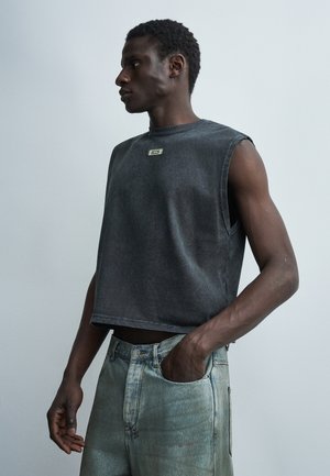 Άντρας φορώντας αμάνικη ξεθωριασμένη μαύρη μπλούζα και ανοιχτόχρωμο μπλε τζιν, στέκεται με το ένα χέρι στην τσέπη, κοιτώντας προς τα αριστερά.