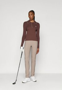 Bruine polo met lange mouwen, een kraag en knoopdetails, gecombineerd met lichtbeige strakke broek en witte sportschoenen, met in de hand een golfclub.