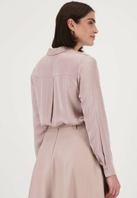 Lichtroze blouse met lange mouwen, kraag en een rugplaid detail. Soepele, zijdeachtige textuur met knoopsluiten aan de manchetten. Zacht, minimalistisch ontwerp.