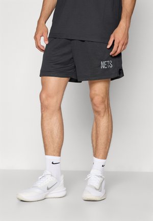 Čierne športové šortky s sieťovinovou textúrou a bočnými rozparkom, s nápisom "NETS" v svetlosivej farbe na ľavom boku. Nosené s bielymi teniskami a ponožkami Nike.