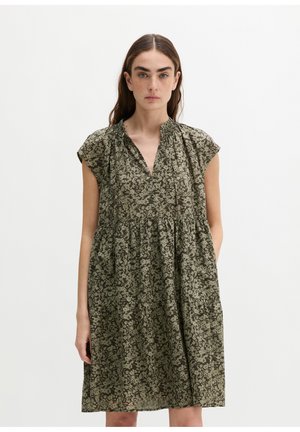 Vrouw met lang bruin haar, die een losse korte mouw jurk draagt met een groen-zwart bloemenpatroon, staand tegen een effen witte achtergrond.