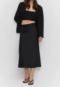 Schwarzes Zweiteiler-Outfit bestehend aus einem taillierten Cropped Top und einem langen, satinierten Rock mit leichtem Glanz. Kombiniert mit schwarzen Flip-Flops.