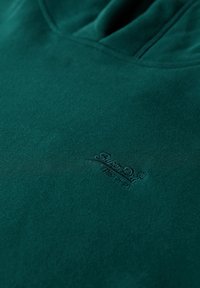 Felpa con cappuccio verde scuro realizzata in un tessuto morbido e testurizzato, con un logo ricamato discreto nell'angolo inferiore.