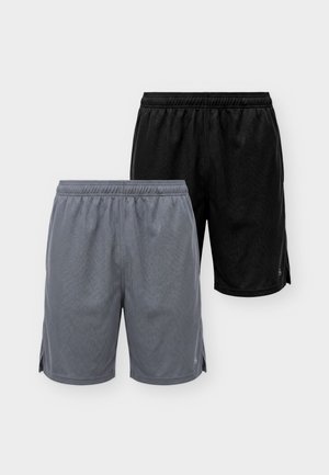 Man draagt een zwart nauwsluitend shirt met lange mouwen, grijze sportshorts, witte sokken en witte sneakers, staand tegen een lichte achtergrond.