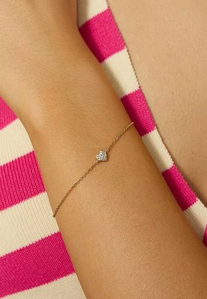 Guldring med en delikat kæde og et lille, hjerteformet charms med en enkelt rhinsten, båret på et håndled.