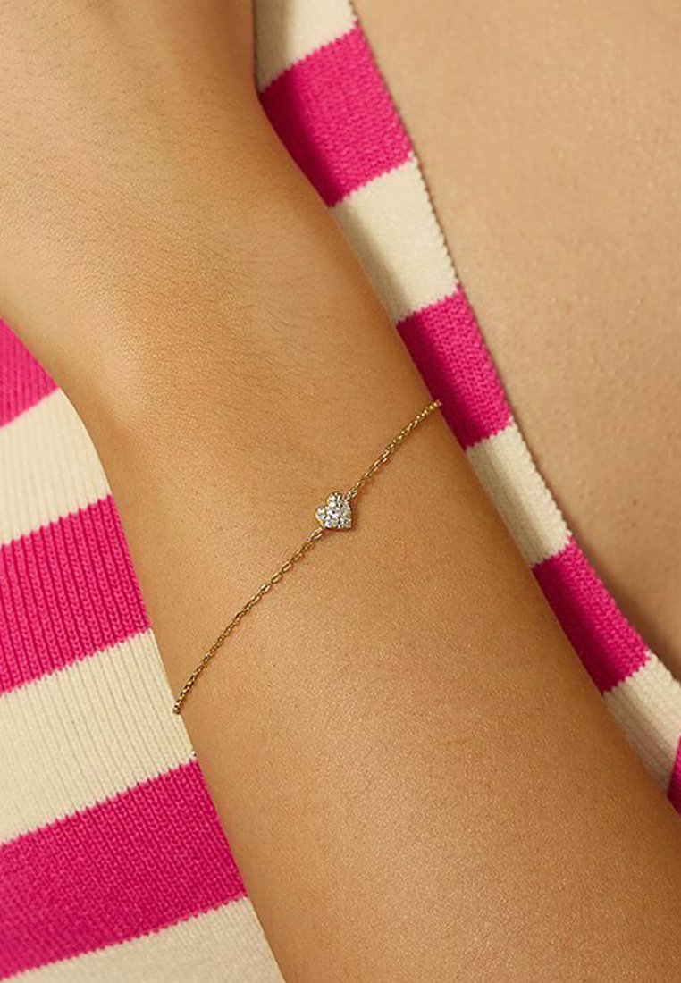 Goldarmband mit einer zarten Kette und einem kleinen, herzförmigen Charm, der einen einzelnen Strassstein trägt, getragen an einem Handgelenk.
