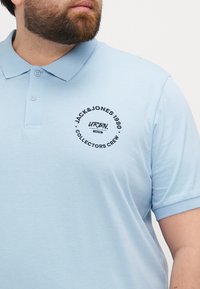 Šviesiai mėlynas polo marškinėliai iš minkštos medvilnės, su apykakle ir trumpomis rankovėmis. Dešinėje krūtinės pusėje yra apvalus juodas logotipas.