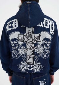 Jeansjacke mit einer blauen Kapuze, die auf der Rückseite ein großes grafisches Motiv eines Kreuzes, Rosen und Totenschädel in Silber zeigt. Ed Hardy-Logo oben.
