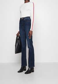 Henkilö, jolla on valkoinen pitkähihainen paita punaisilla raidoilla, tummansininen korkeavyötäröiset farkut, mustat nilkkurit ja joka pitää mustaa nahkalaukku.