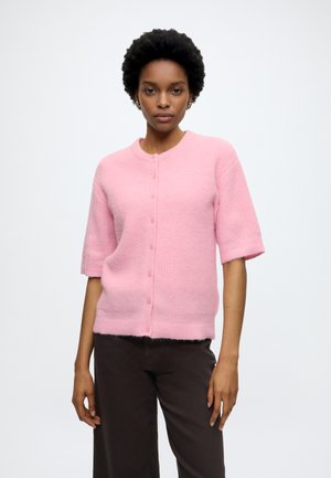 Femme portant un cardigan court à manches courtes rose pâle boutonné et un pantalon sombre, debout devant un fond clair uni.