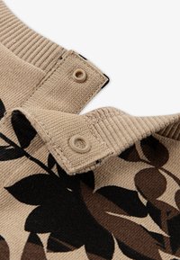 Beige strikket stoff med svart og brunt blomstermønster. Har en ribbestrikket krage og to knapper ved åbningen. Strukturert finish.