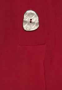 Roter Stoff mit glatter Textur zeigt einen strukturierten, unregelmäßig geformten Knopf aus hellfarbigem Perlmutt mit einem zentralen Loch.