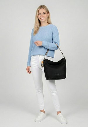 Young woman in light blue sweater, white pants, and sneakers holding a black shoulder bag with decorative keychains, smiling at camera.