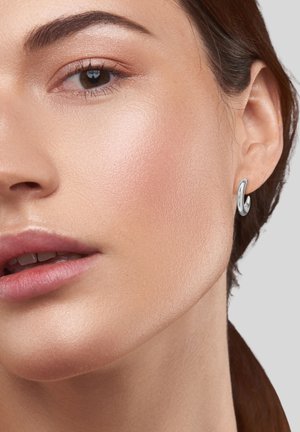 Boucles d'oreilles en argent au design courbé et élégant, fixées à un lobe d'oreille, mettant en valeur une peau lisse et un maquillage naturel.