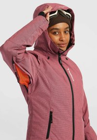 Persoon in een kastanjebruine winterjas met capuchon, voorzien van ventilatieopeningen op de mouwen, en witte skibril omhoog op het hoofd.