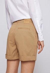 Person som bär beige uppvikta shorts med en BOSS-etikett på midjan och en vit långärmad skjorta instoppad.