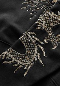 Sudadera negra con intrincados adornos en oro y plata, incluyendo cuentas y lentejuelas dispuestas en un diseño estilizado.