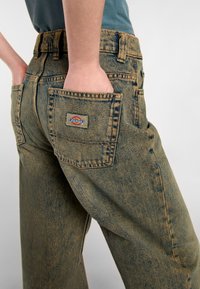 Denimbyxor i blekt olivgrönt, med hög midja, lös passform och bakfickor. Dickies-logotyp på höger ficka.