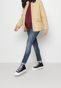 Chaqueta acolchada beige con cuello de piel, combinada con una blusa burdeos y jeans ajustados azules. Zapatillas altas negras completan el conjunto.
