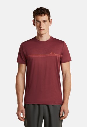 Mann mit kurzen lockigen Haaren, der ein weinrotes T-Shirt mit einer dünnen orangen Linie und einem geometrischen Muster über der Brust sowie dunkle Hosen trägt.