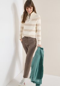 Femme portant un pull beige à rayures et un pantalon marron, tenant une veste en velours côtelé bleu sarcelle, souriant et regardant vers le bas sur un fond uni.