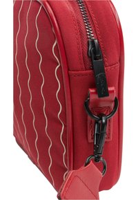 Červená crossbody taška z látky s vlnitým bílým vzorem, černým zipem a hardwarovými detaily, která se vyznačuje elegantním designem a texturovaným povrchem.