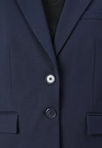 Navy blazer van soepel materiaal, met twee knopen, geklede revers en twee voorzakken. De knopen zijn zwart met een gestructureerd ontwerp.